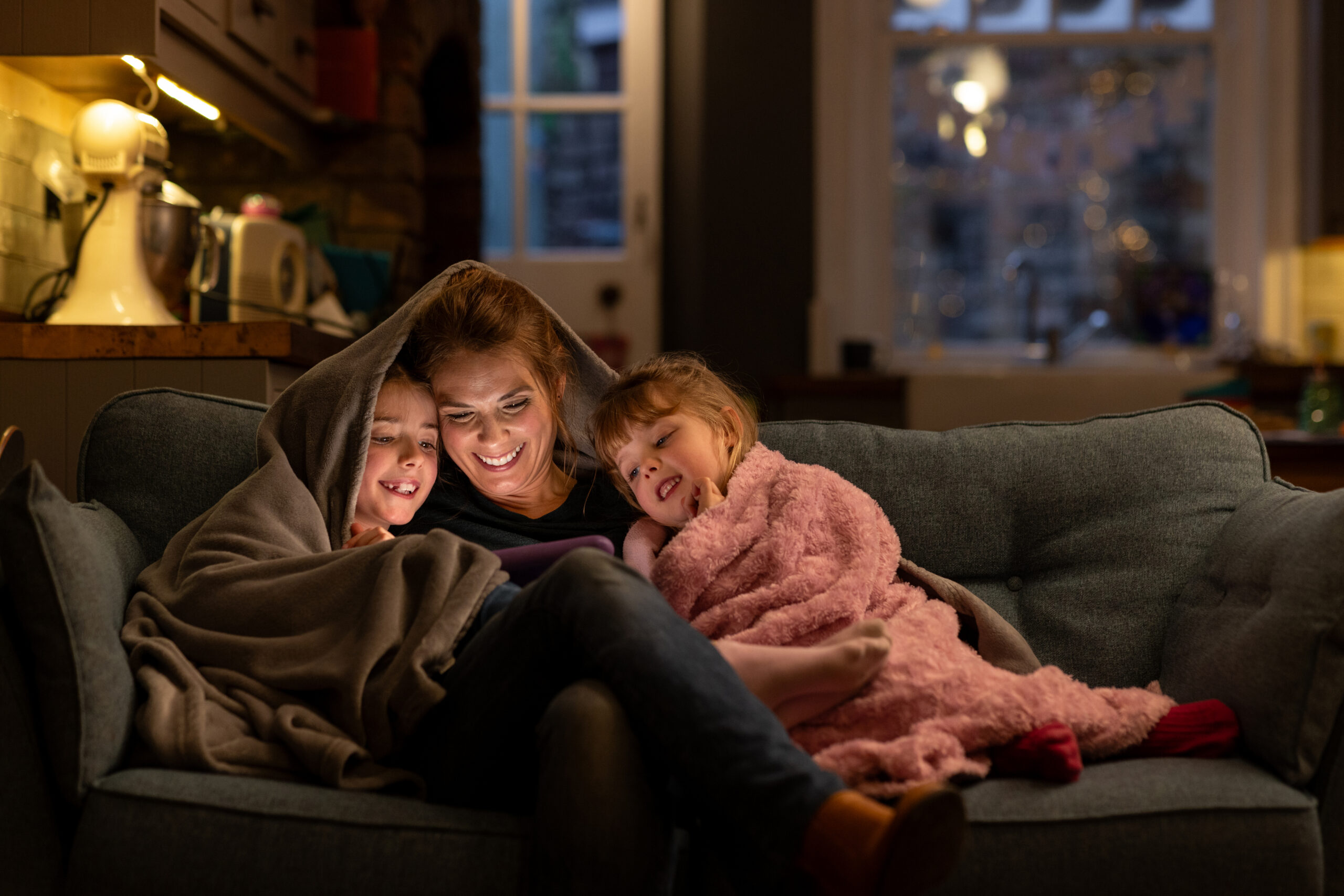 A shot of two young girls with their mother, sitting comfortably with a blanket over their heads on a sofa in the living room of their home in South Shields, North East England. They are all smiling, looking at a digital tablet which the mother is holding. In the background the kitchen is in partial view, and it is dusk outside.

Video similar to this scenario available.